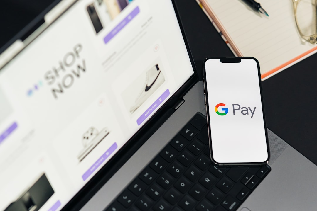 Google Pay displayed on a phone screen. Top-down view of a workspace. Desk table with a laptop showing ecommerce on screen, a mobile phone, and a notepad
